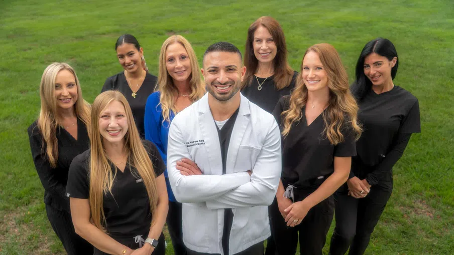 Smiling dental team with male dentist in white coat and seven female staff on green grass background