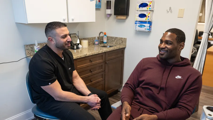A dentist in black scrubs talking to a male patient in a maroon Nike tracksuit in a dental office.
