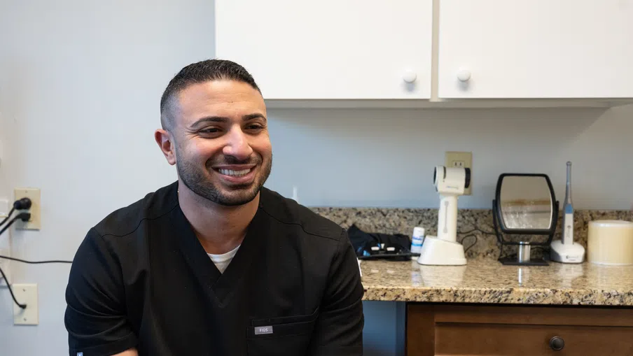 Smiling healthcare professional in black scrubs sitting in a medical office with equipment in the background