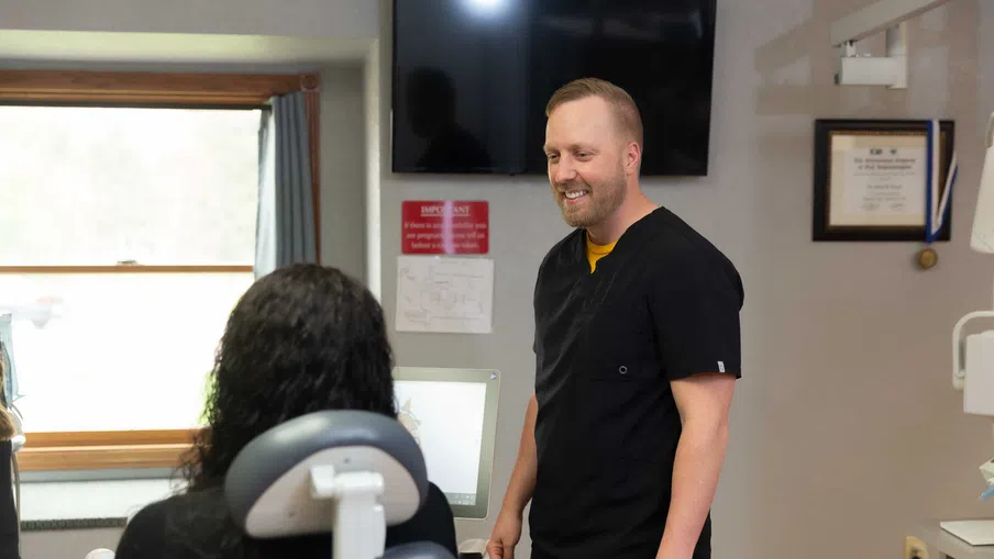 Dentist in black scrubs talking to a patient seated in a dental chair in a clinic room with medical equipment.