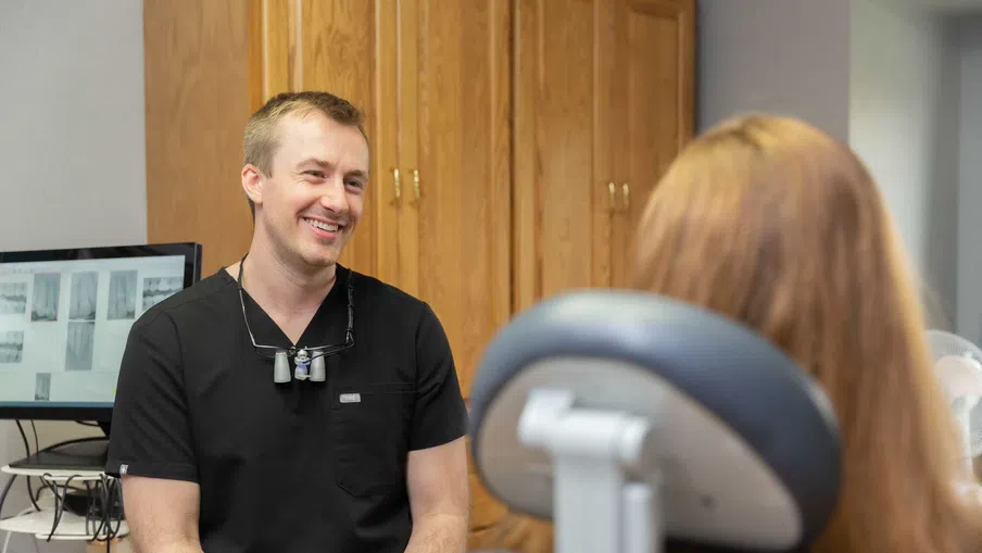 Smiling male dentist in black scrubs talking to a female patient in a dental office with wooden cabinets.