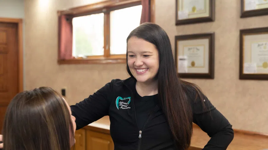 Smiling female dental professional interacting with a patient in a warmly lit dental office with framed certificates.