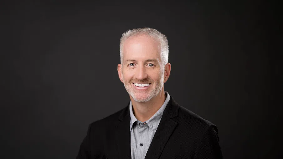 Smiling middle-aged man with gray hair and beard wearing a black blazer and gray shirt on dark background