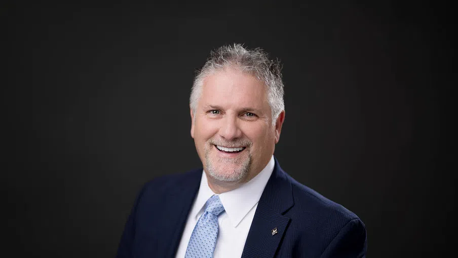 Smiling middle-aged man in navy suit and light blue tie against a dark background professional headshot.