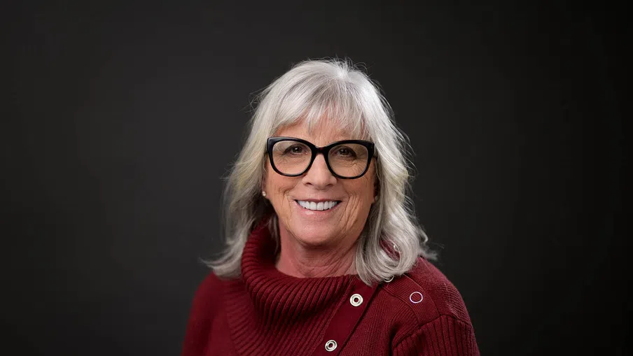 Smiling older woman with gray hair and black glasses wearing a red sweater against a dark background