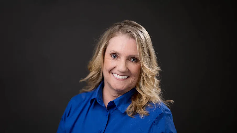 Smiling middle-aged woman with wavy blonde hair wearing a bright blue button-up shirt against a dark background