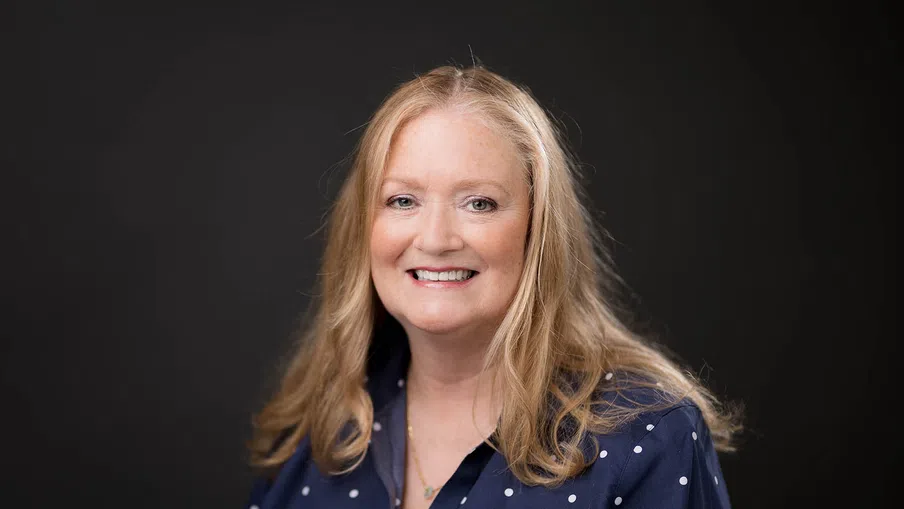 Smiling middle-aged woman with long blonde hair wearing a navy blue polka dot blouse against a dark background