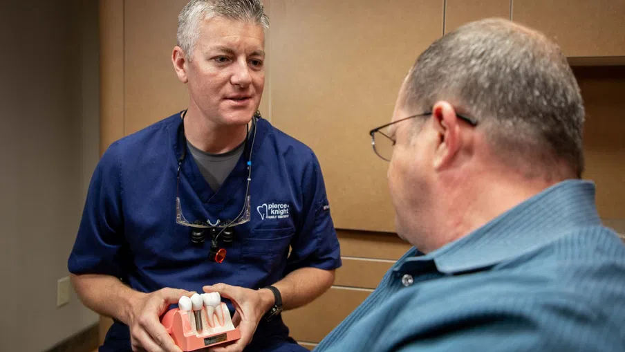 Dentist in blue scrubs explaining dental implant model to male patient in office consultation.