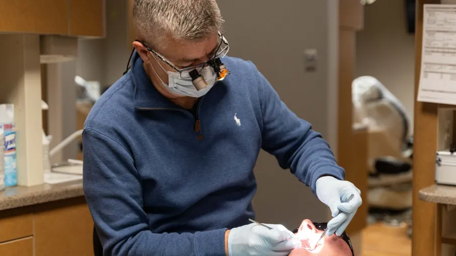 Dentist wearing magnifying glasses and gloves examining a patient's teeth in a dental clinic.