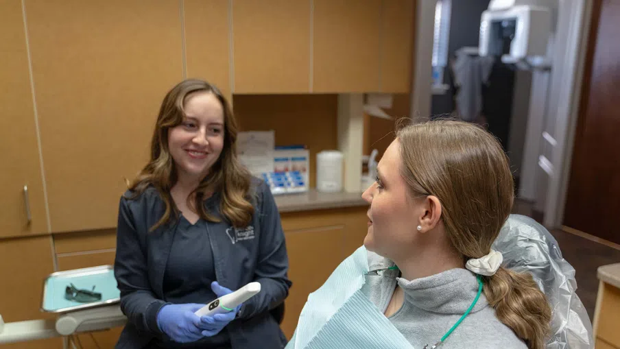 Dentist smiling and holding a dental tool while talking to a patient seated in a dental chair with a bib.