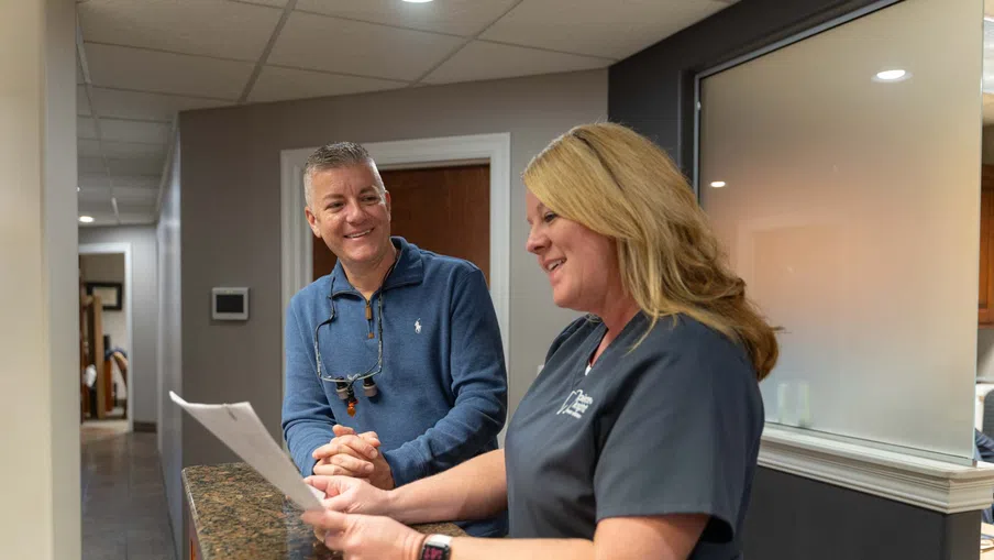 Dental professional discussing documents with a smiling male patient at the reception desk in a modern clinic.