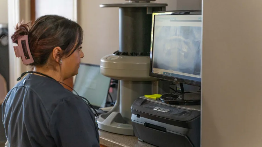 Woman in a dental office viewing dental X-rays on a computer screen next to imaging equipment.