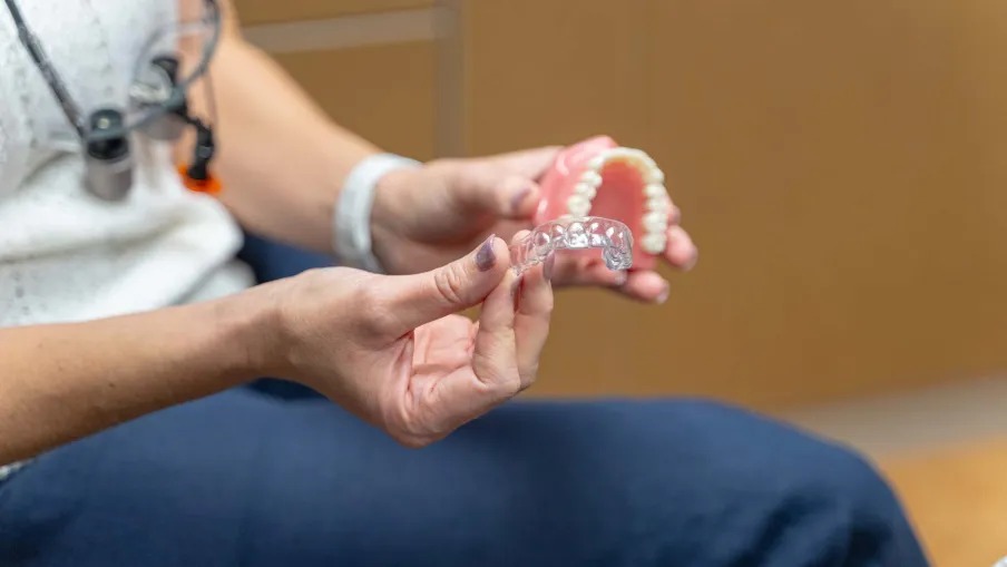 Person holding clear dental aligner and pink denture model in a dental office setting