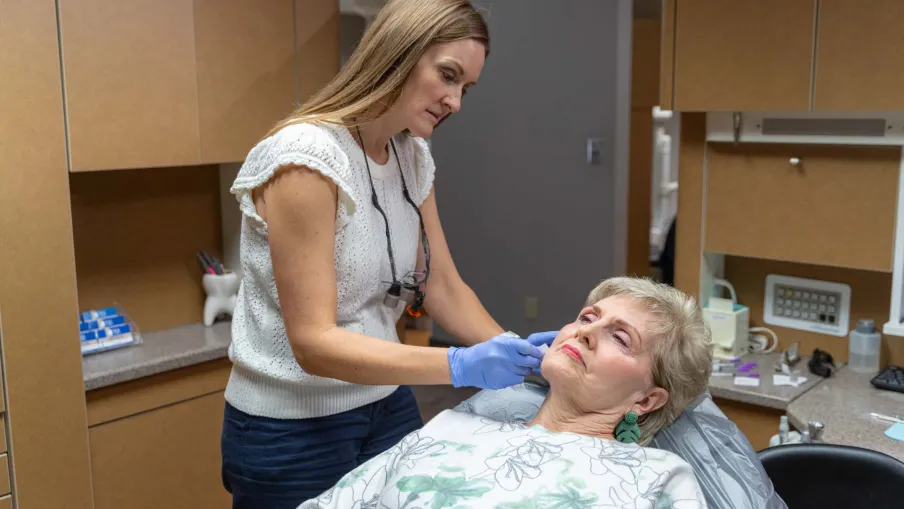 Dentist examining elderly woman's teeth in a dental clinic setting with medical tools and cabinets.