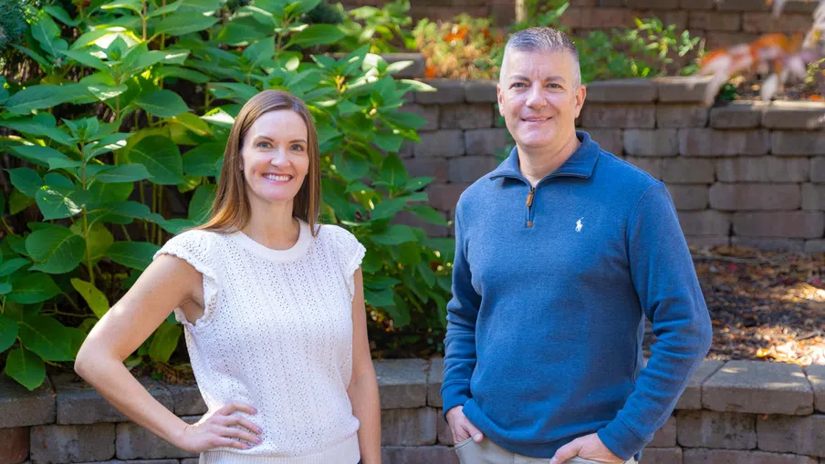 Two adults smiling outdoors by stone wall and green plants, woman in white sleeveless top and man in blue sweater.