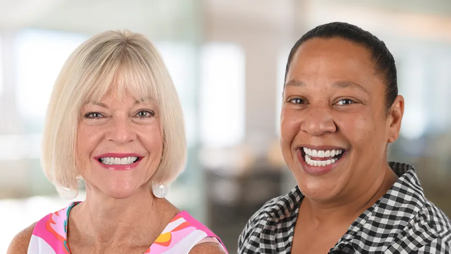 Two smiling women, one with short blonde hair in a colorful dress, the other with dark hair in a checkered shirt.