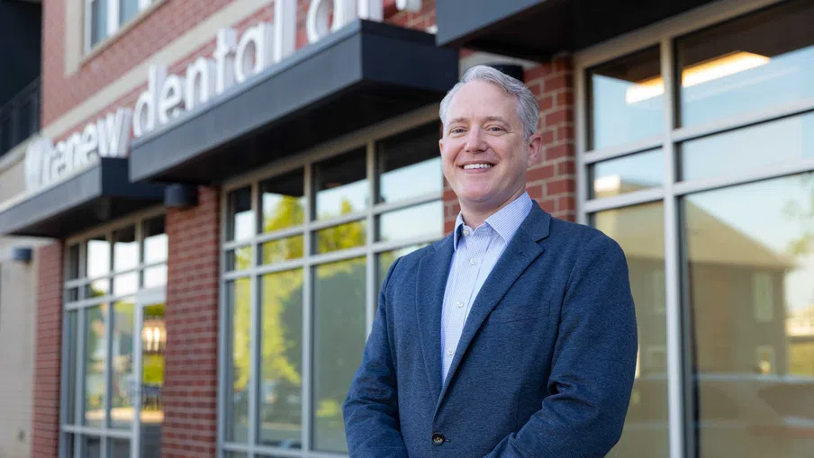 Smiling man in blue blazer standing outside a Modern dental office with glass windows and brick walls
