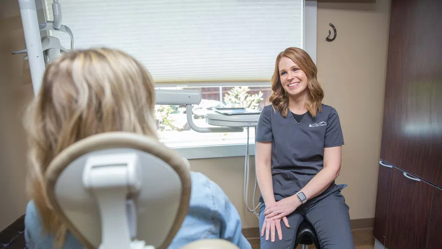 Dental professional in gray scrubs consulting with patient in bright dental office.