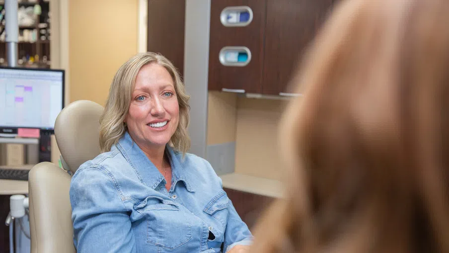 Smiling woman in denim shirt sitting in a dental office chair during a consultation with a dentist.