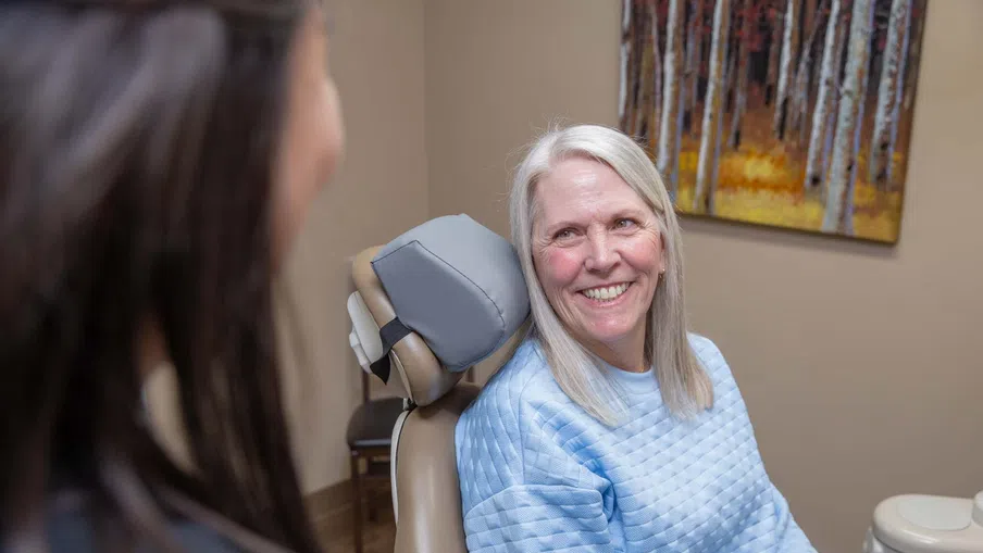 Smiling elderly woman in a blue sweater sitting in a dental chair talking to a dentist.