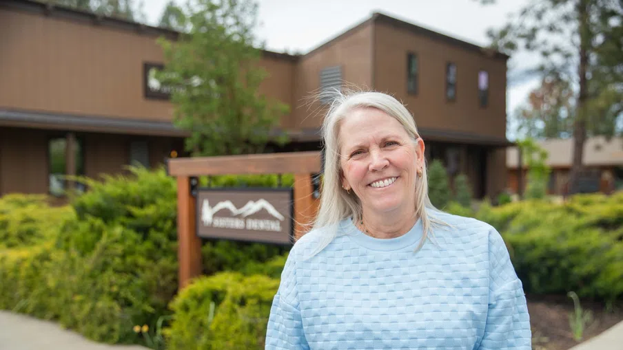 Smiling older woman in light blue sweater standing outside a dental office with greenery and wooden sign.
