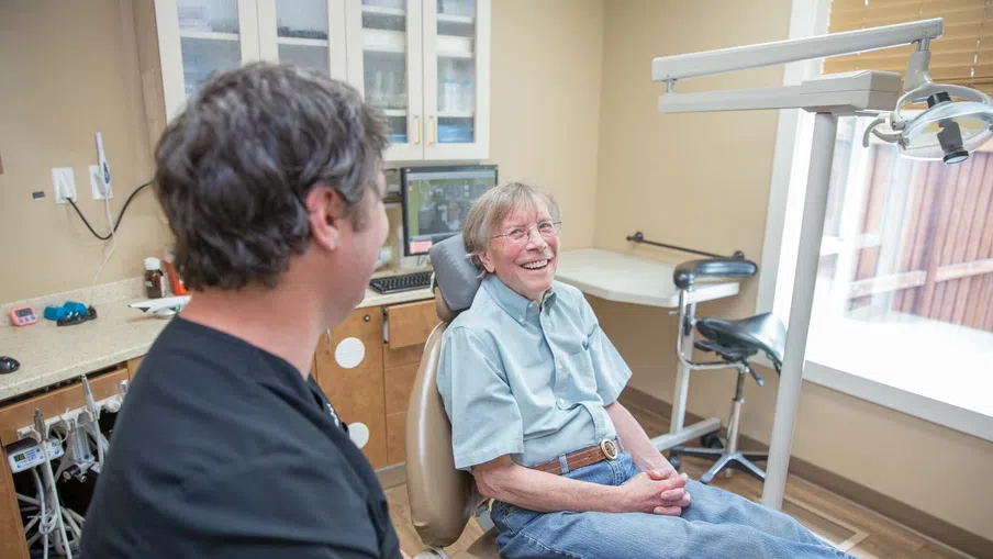 Elderly man smiling while sitting in dental chair talking to dentist in a bright clinic room