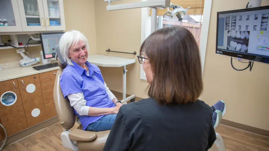 Elderly woman sitting and smiling in dental chair during consultation with female dentist in clinic.