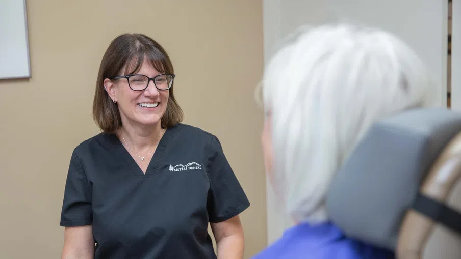 Smiling female dental professional in black scrubs talking to elderly patient in dental chair.