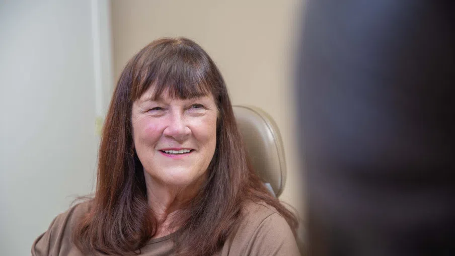 Smiling middle-aged woman with brown hair sitting in an office chair wearing a brown top.