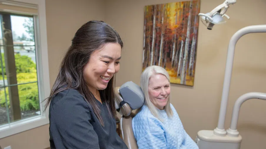 Smiling female dentist in black scrubs with elderly female patient in dental chair in bright clinic room