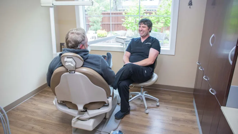 Dentist and patient having a friendly conversation in a bright dental office with wooden floors and large window.