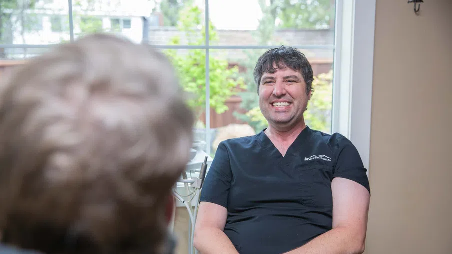 Smiling male healthcare professional in black scrubs sitting and talking with a patient in a bright room.