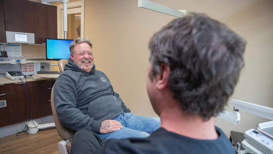 Smiling man in hoodie sitting in dental chair talking to dentist in clinic with medical equipment.