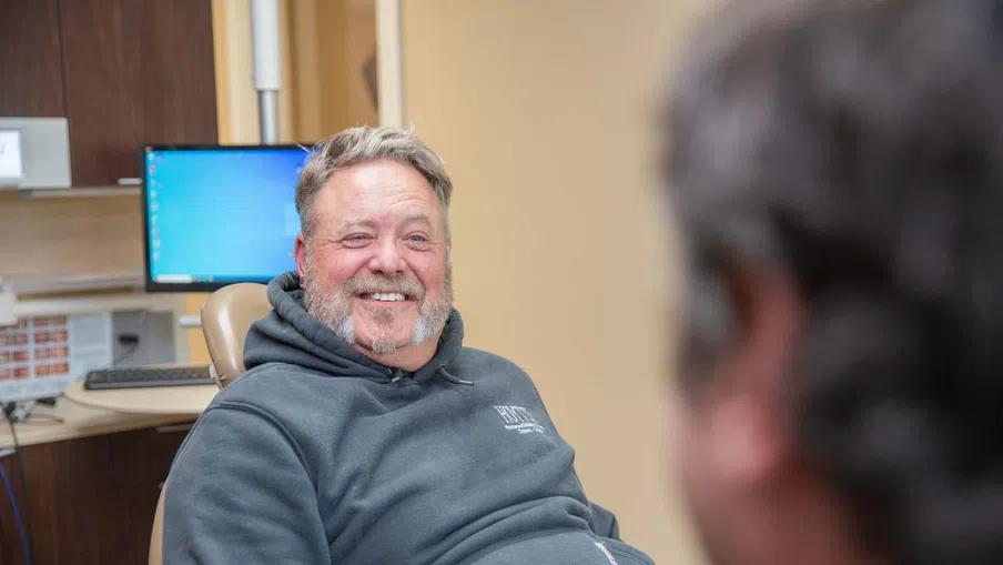 Smiling man in a gray hoodie sitting in an office chair with a computer in the background.