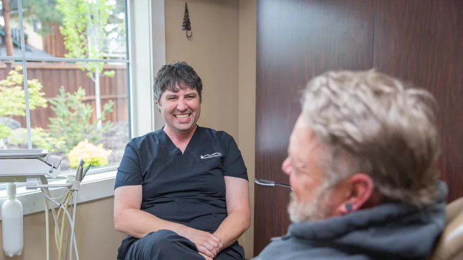 Smiling male dentist in black scrubs talking to relaxed male patient in dental office with window view.
