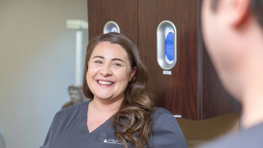 Smiling female healthcare worker in gray scrubs talking to a colleague in a medical office setting.