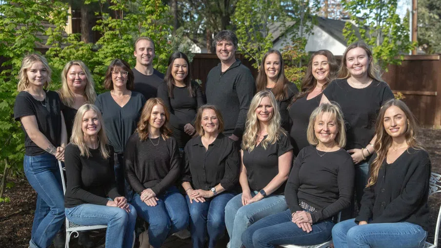 Group of fifteen diverse adults wearing black tops and jeans, posing outdoors on a sunny day with greenery and a patio.
