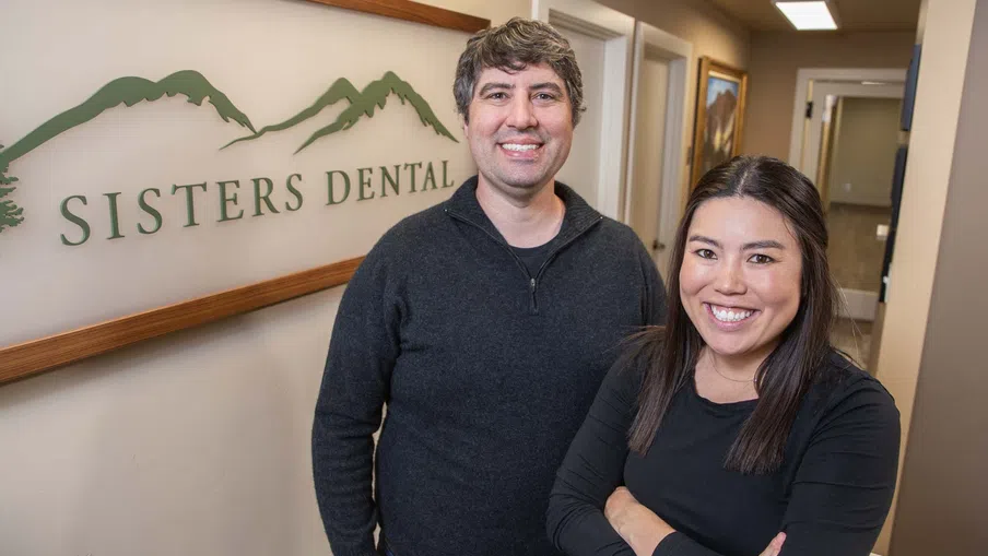 Two dental professionals smiling inside Sisters Dental office with logo and mountain design on wall.
