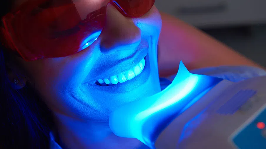 Woman wearing red protective glasses undergoing blue light teeth whitening treatment at dental clinic.