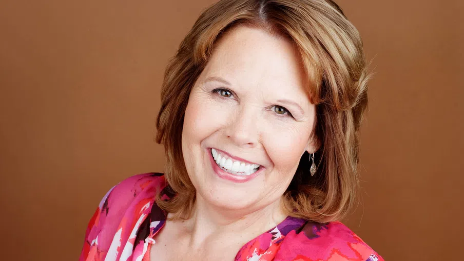 Smiling middle-aged woman with short light brown hair wearing a bright floral blouse against a brown background