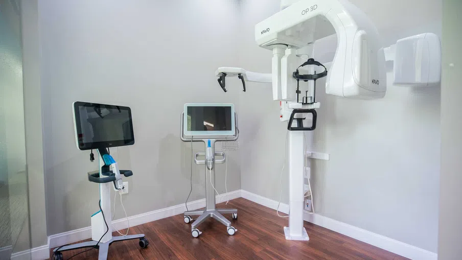 Dental imaging room with a 3D panoramic X-ray machine and two computer monitors on mobile stands.