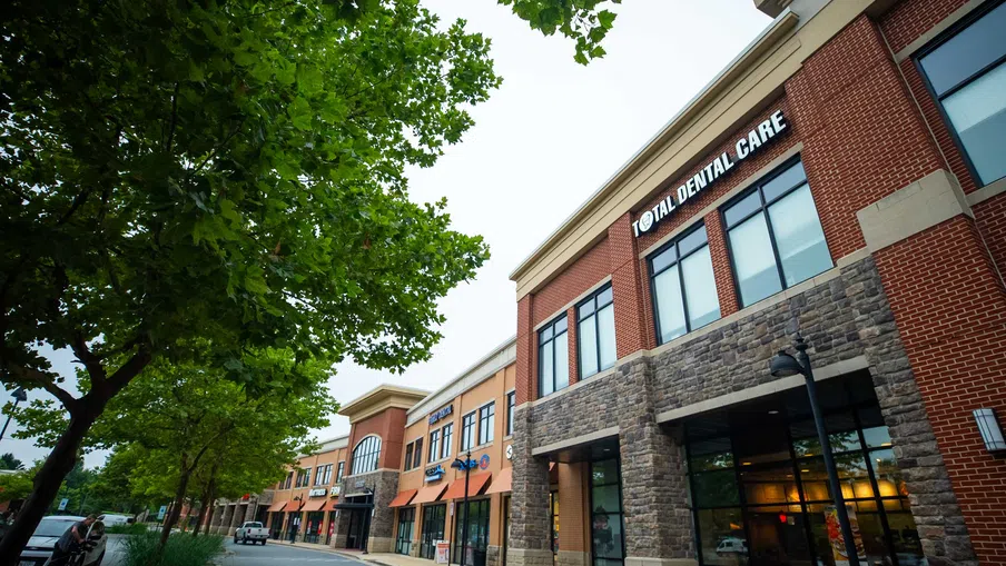 Modern commercial building with Total Dental Care and other storefronts under green trees on a cloudy day.