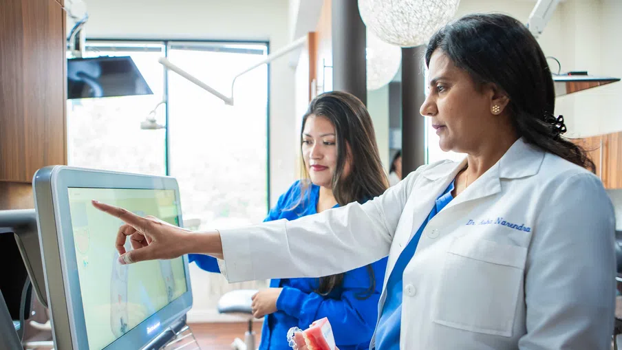Female dentist in white coat showing dental model and pointing at screen with assistant in clinic.