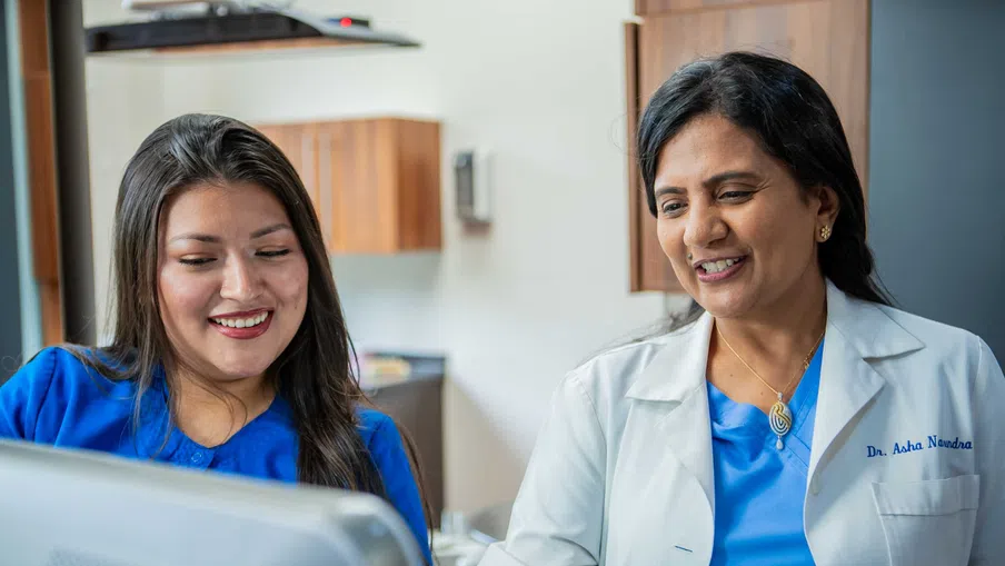 Two female healthcare professionals in blue scrubs reviewing information on a computer screen in a medical office.