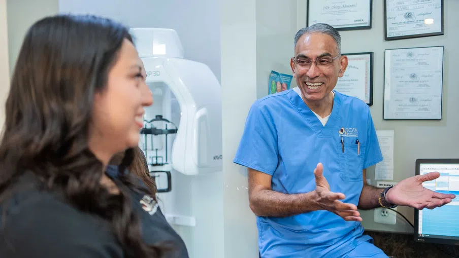 Smiling male dentist in blue scrubs talking with female patient in a dental office with equipment and certificates
