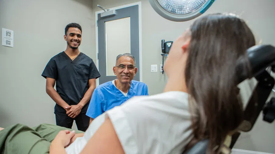 Two healthcare professionals smiling at a patient reclining in a medical examination chair in a clinic room.
