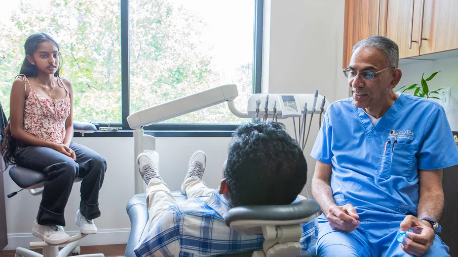 Dentist in blue scrubs talking to a male patient in a dental chair while a young girl waits nearby.