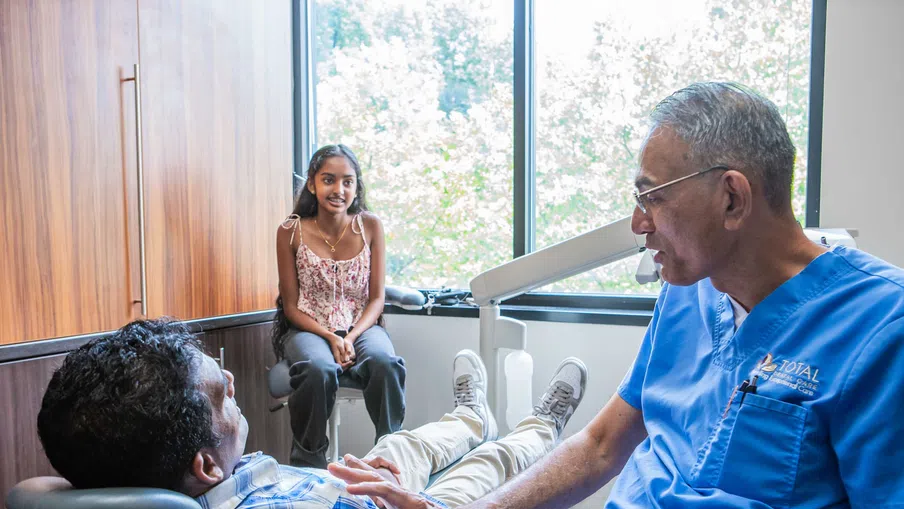 Dentist in blue scrubs talking to male patient in dental chair while young girl sits nearby by window.