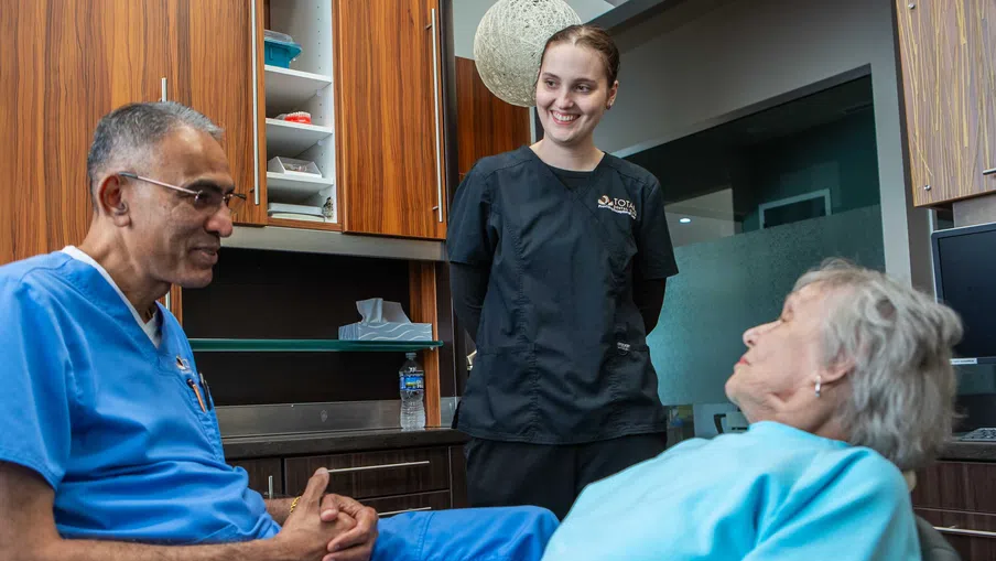 Dentist in blue scrubs talking to elderly patient in chair with nurse standing and smiling in dental office.