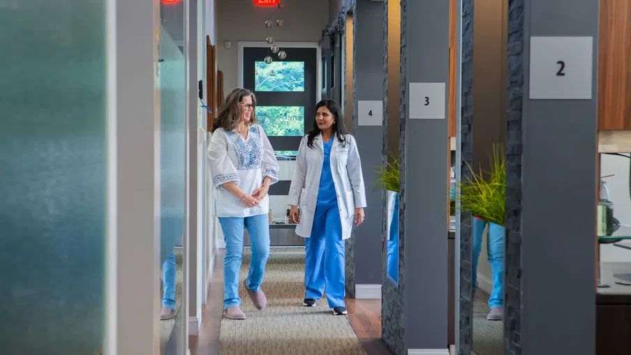 Doctor and patient walking and talking together in a modern medical clinic hallway with numbered rooms.